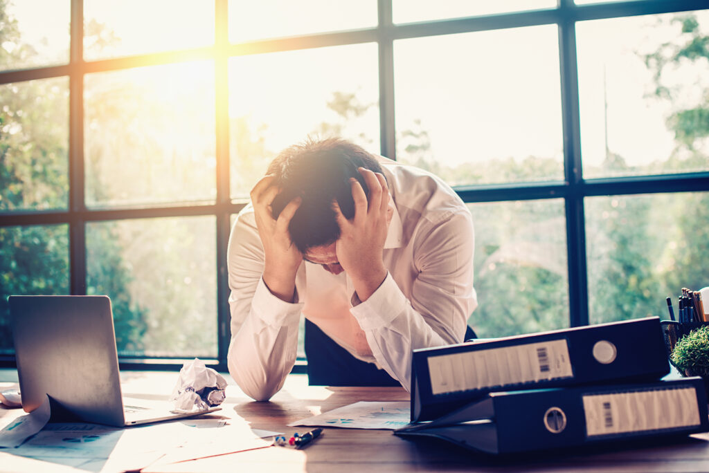 Man sitting at a cluttered desk, hands on head in frustration, sunlight from large windows behind him.