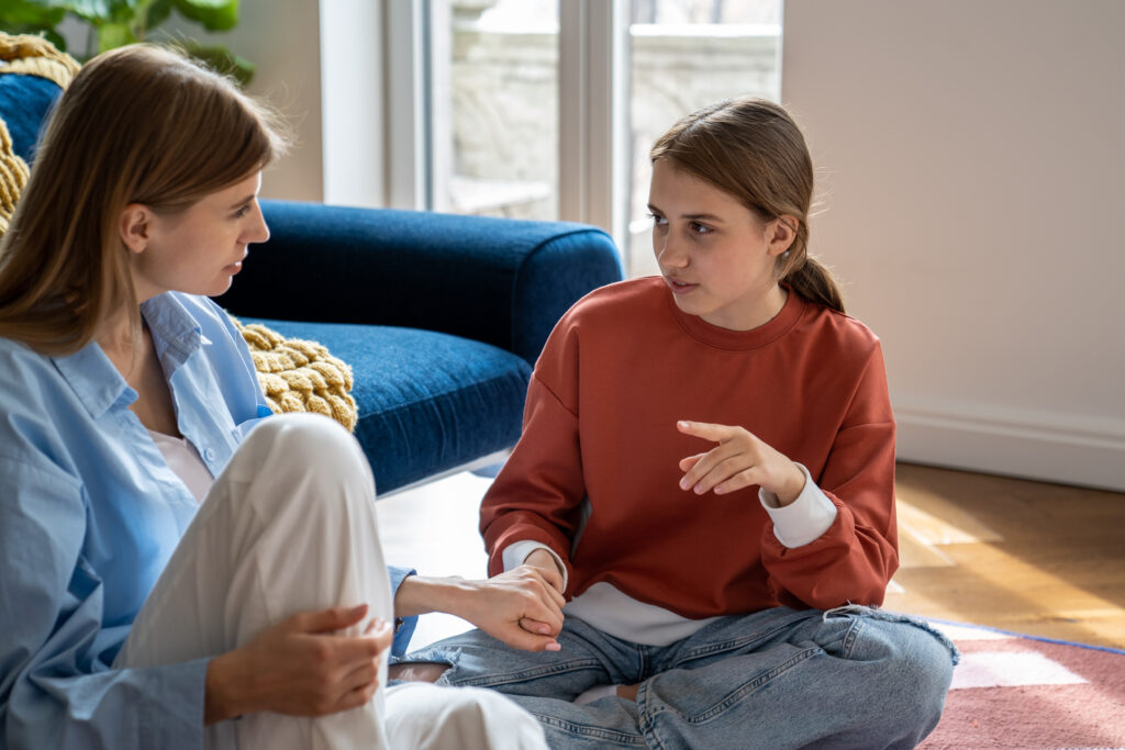Adult and child sit cross‑legged on the floor, talking warmly in a living room by a blue sofa and window light.