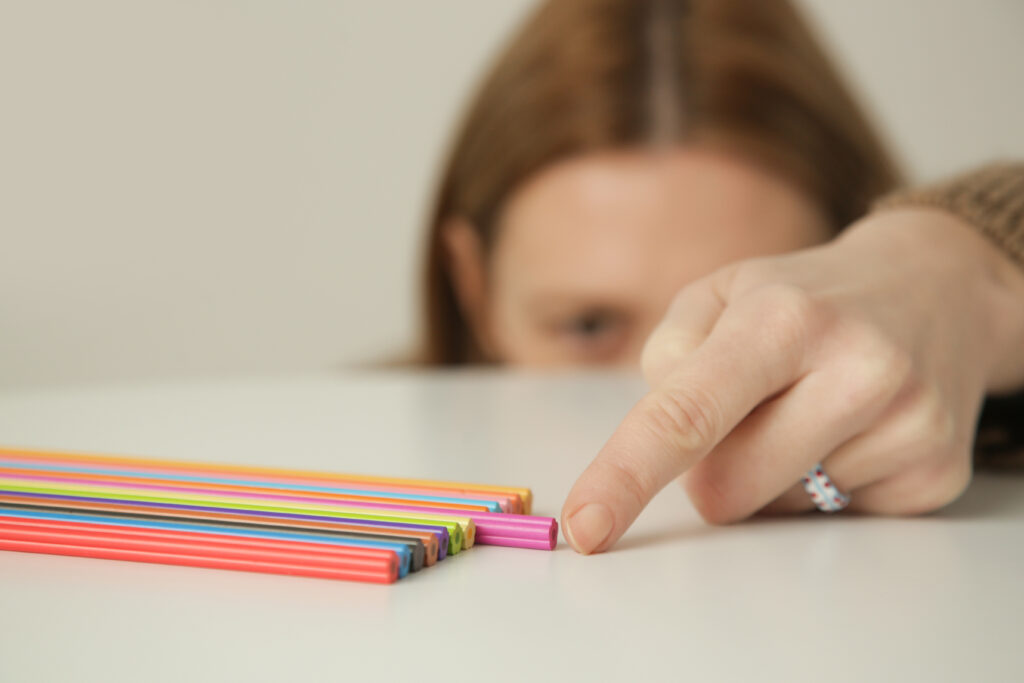A hand reaching toward a row of brightly colored pencils lined up on a white table.