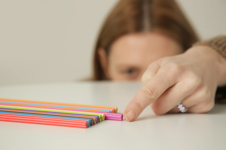 A hand reaching toward a row of brightly colored pencils lined up on a white table.