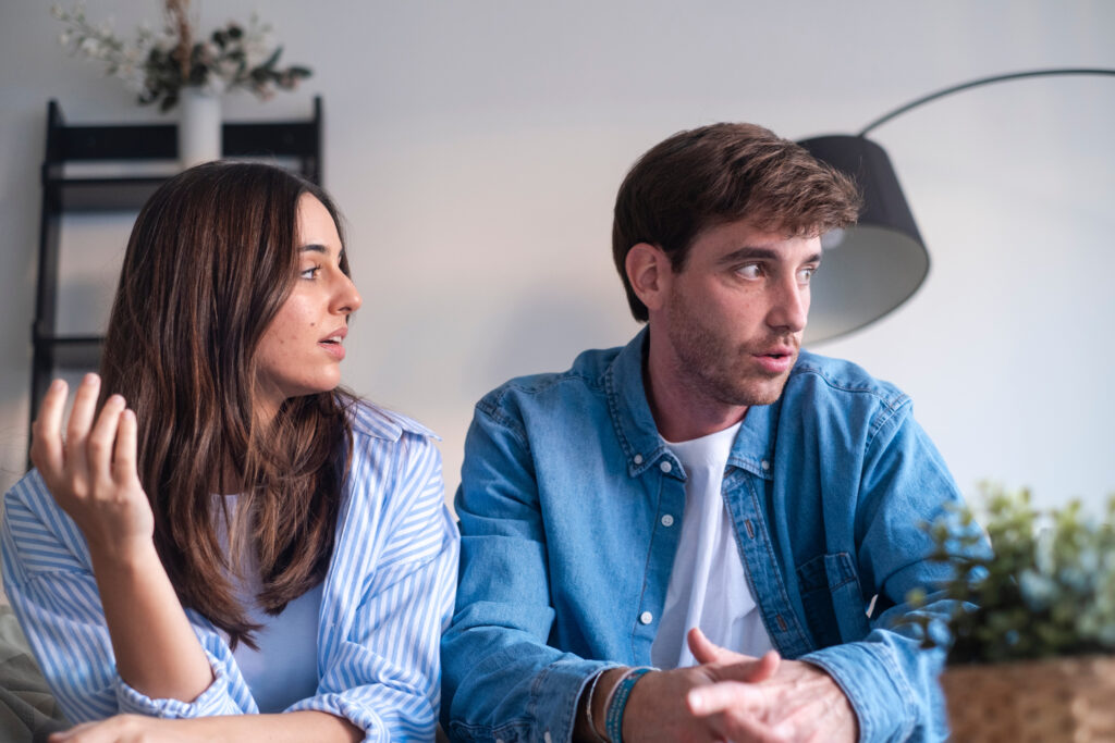 Two young adults sit and talk in a bright room; woman on the left gestures while the man on the right listens.