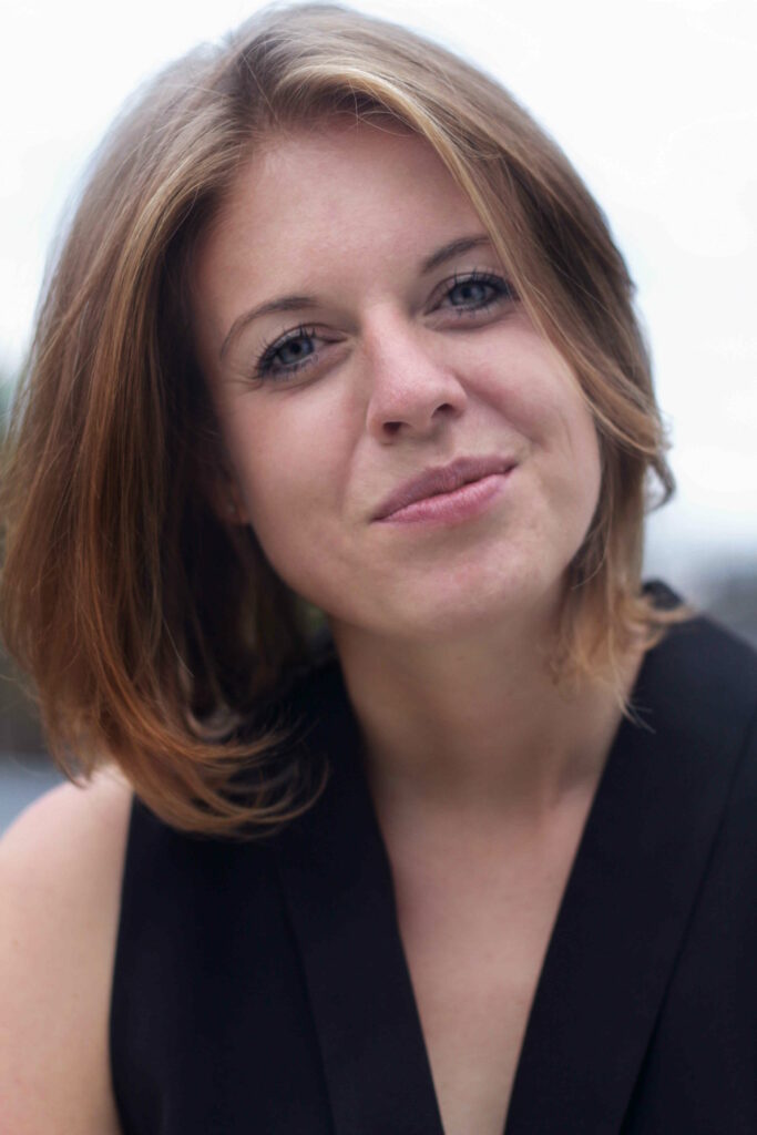 Portrait of a woman with short light brown hair smiling softly, wearing a black sleeveless top outdoors.